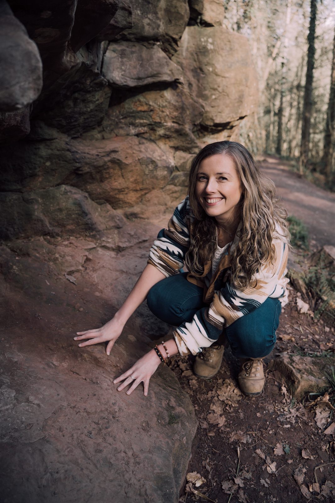 Libby smiling, crouched with both hands on a red sandstone ledge beside a woodland path